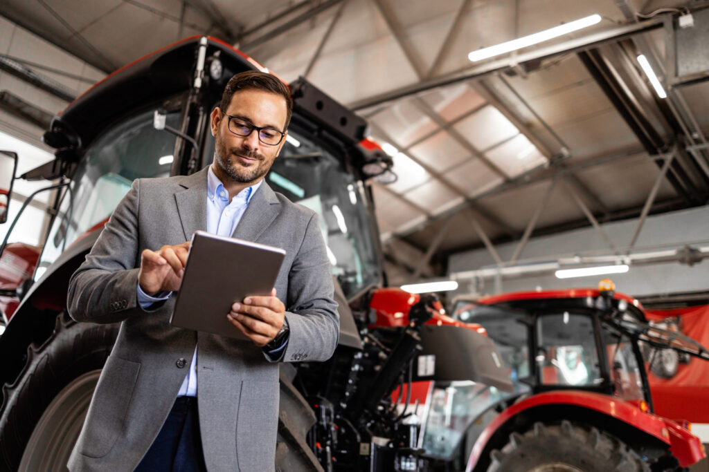 Experienced Salesman Standing By Tractor Machines At Dealership And Checking Agricultural Equipment On Tablet Computer.
